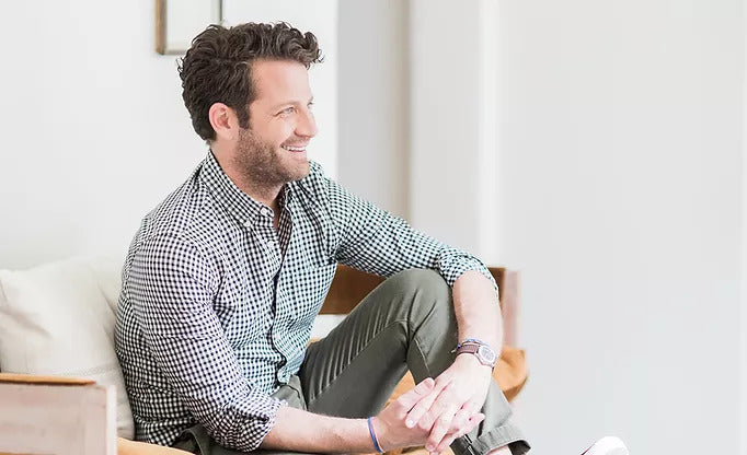 Nate Berkus sits in a chair, turned slightly away from the camera and smiling. 
