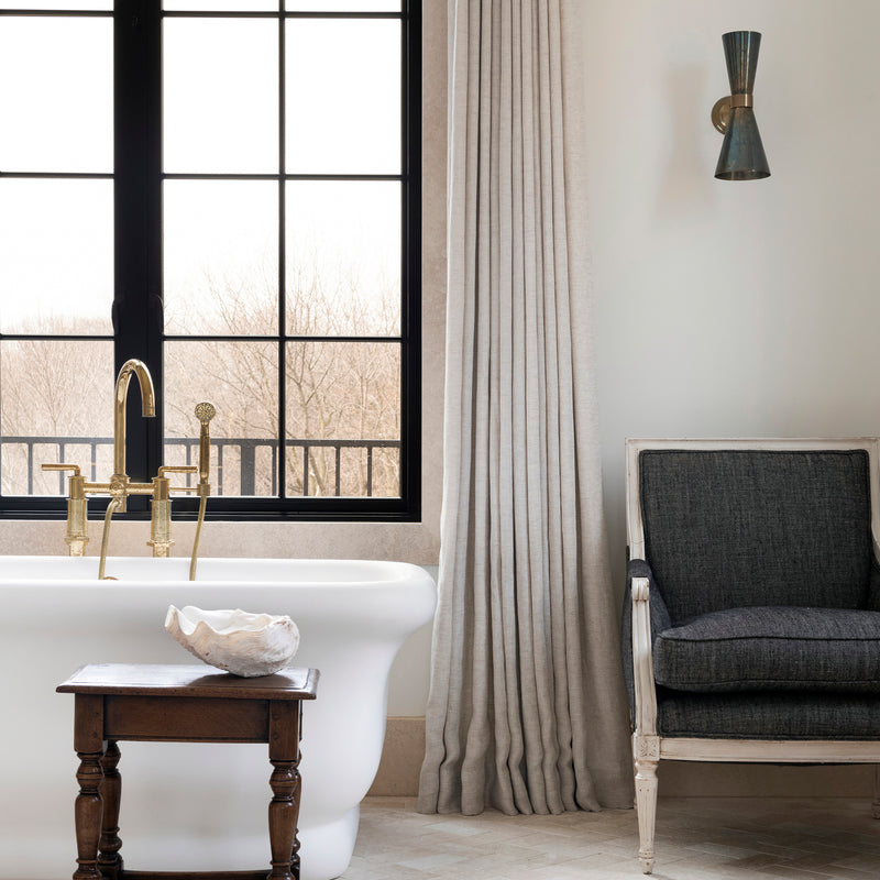 A Nate Berkus Associates bathroom with a soaking tub. The room features black windows, modern sconces, and antique furnishings.