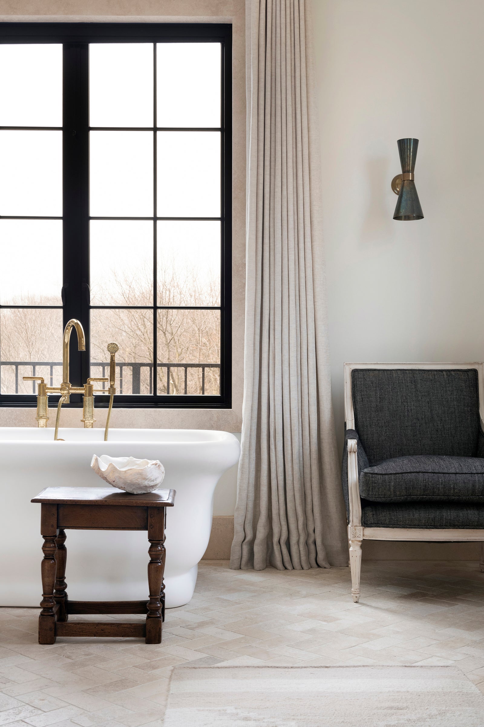 A Nate Berkus Associates bathroom with a soaking tub. The room features black windows, modern sconces, and antique furnishings.
