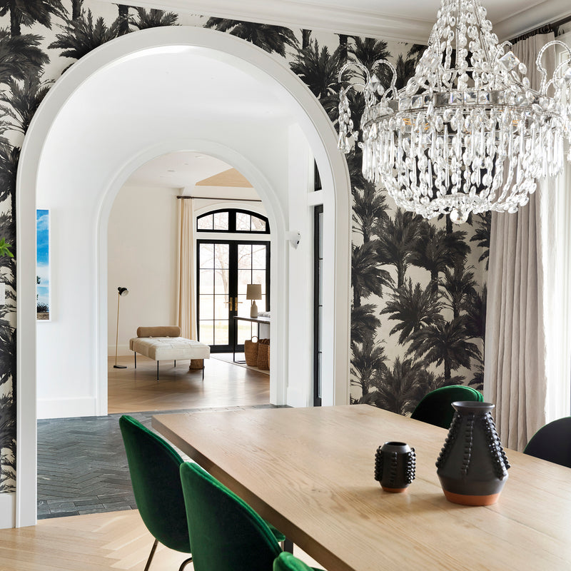 A dining room by Nate Berkus Associates with palm tree wallpaper, herringbone floors, a crystal chandelier, and an arched entry.
