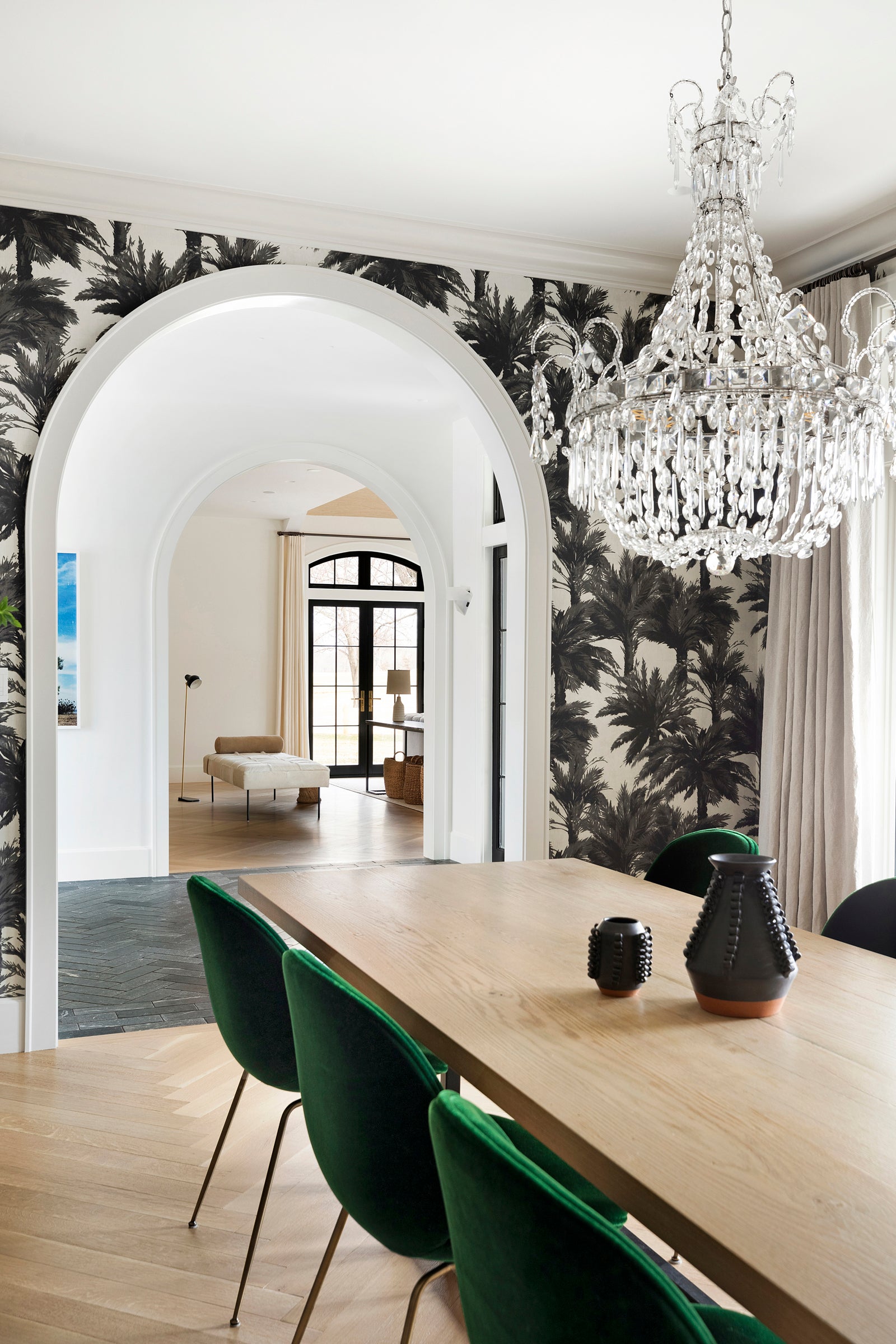 A dining room by Nate Berkus Associates with palm tree wallpaper, herringbone floors, a crystal chandelier, and an arched entry.