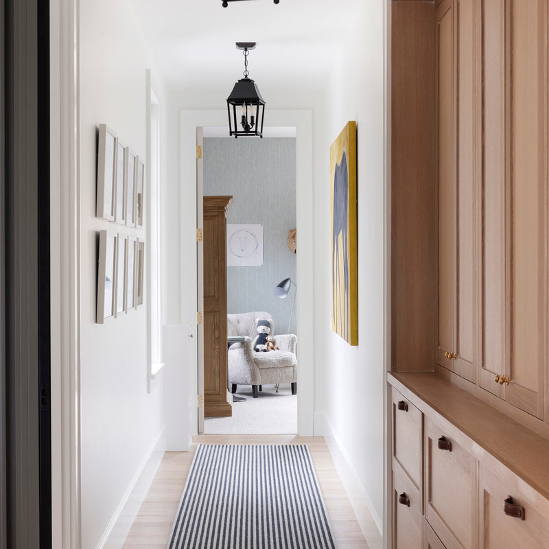 A hallway by Nate Berkus Associates with a striped rug, lantern pendants, statement art, and built-in wood-toned cabinetry.