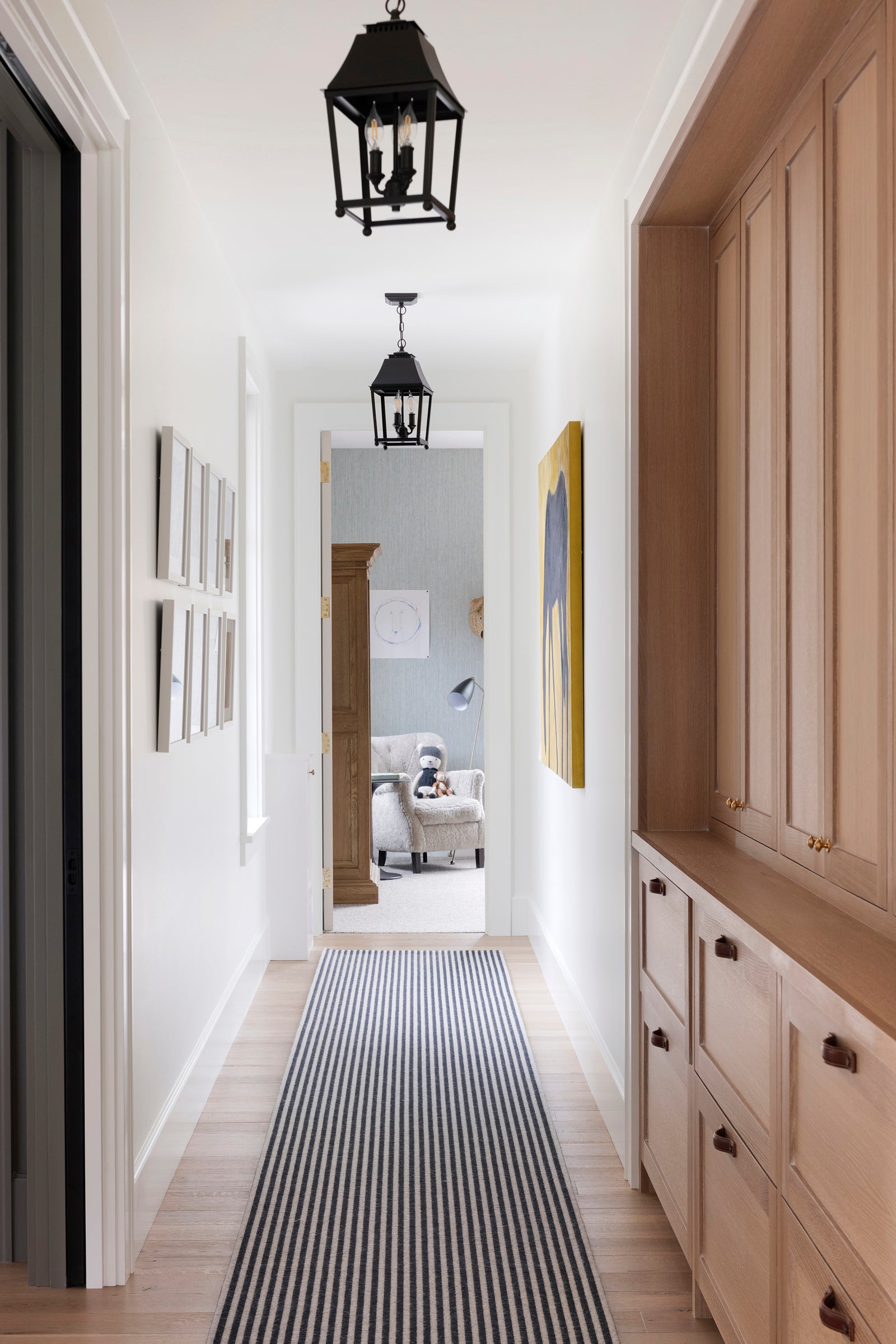 A hallway by Nate Berkus Associates with a striped rug, lantern pendants, statement art, and built-in wood-toned cabinetry.