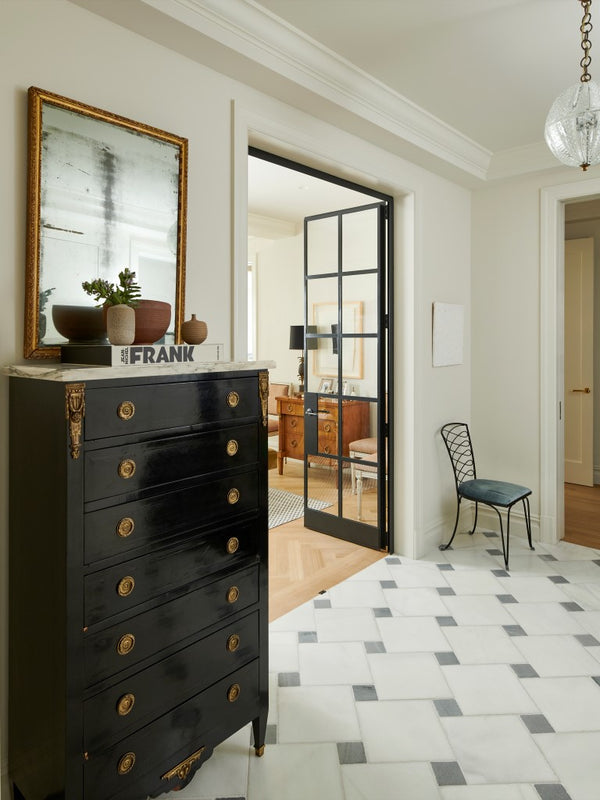 A foyer featuring a checker stone floor, a vintage black lacquer chest with a gilt mirror and an iron and glass door. 