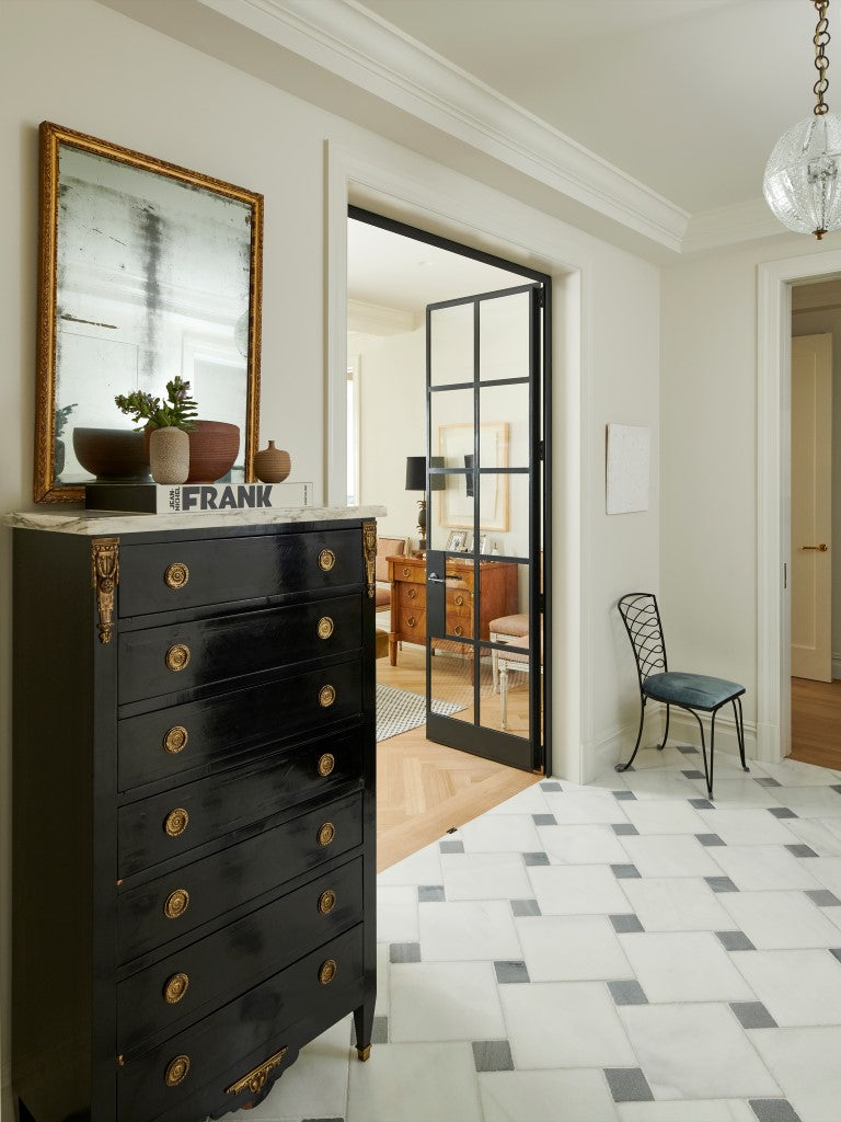 A foyer featuring a checker stone floor, a vintage black lacquer chest with a gilt mirror and an iron and glass door. 