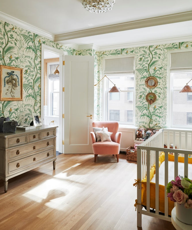 A child's nursery with green botanical wallpaper, a grey painted dresser, crib, and pink upholstered armchair over wood floors.