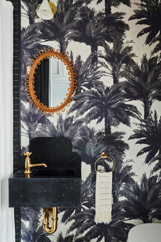 Close up on a powder room wall with a black marble sink with brass fittings, black palm tree wallpaper and a vintage French rattan oval mirror.
