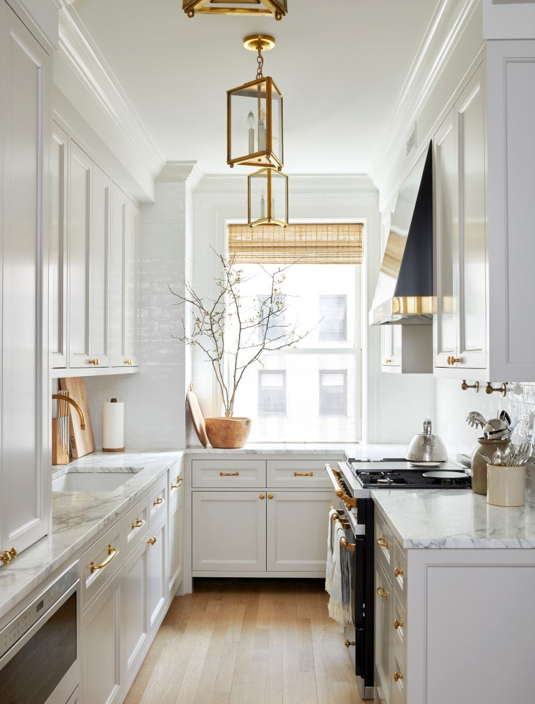 Kitchen with pale grey painted cabinets, brass lighting, and branches in a pot on the windowsill. 