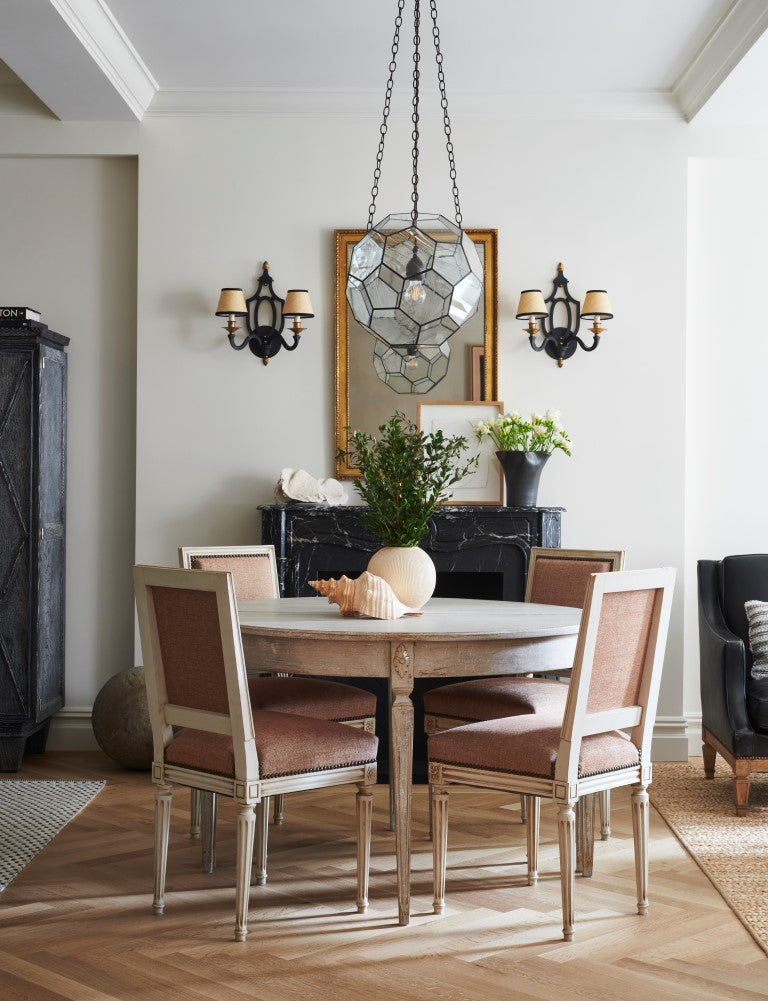 A living room featuring four vintage French chairs, a round oak dining table, a pair of vintage sconces flanking a gilt mirror. An iron and glass pendant hangs over the table.