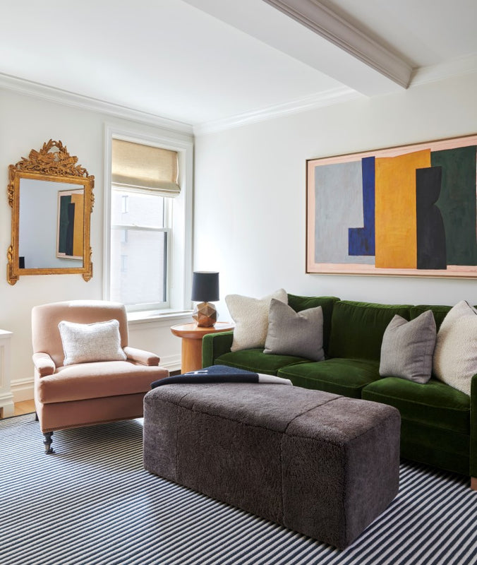 Family room with a green upholstered sofa, a blush velvet armchair, and a grey shearling rectangular ottoman. Colorful abstract art and a gilt mirror hang on the walls. 
