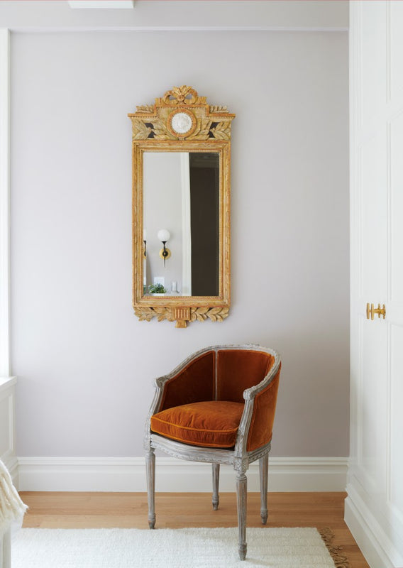 A vignette showing an antique French painted armchair with burnt orange upholstery and an ornate carved gilt mirror on the wall behind it.