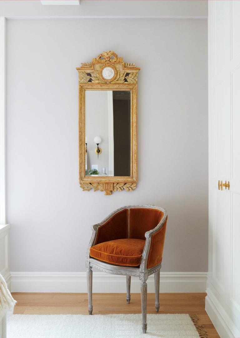 A vignette showing an antique French painted armchair with burnt orange upholstery and an ornate carved gilt mirror on the wall behind it.