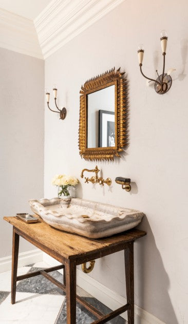 A bathroom sink scene with a natural stone sink bowl, antique table base, and candle sconces. Design by Nate Berkus Associates and Jeremiah Brent.