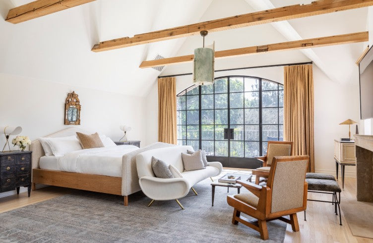 A primary bedroom by Nate Berkus Associates and Jeremiah Brent with rustic wood beams, modern black windows, and a curation of neutral-toned furnishings. 