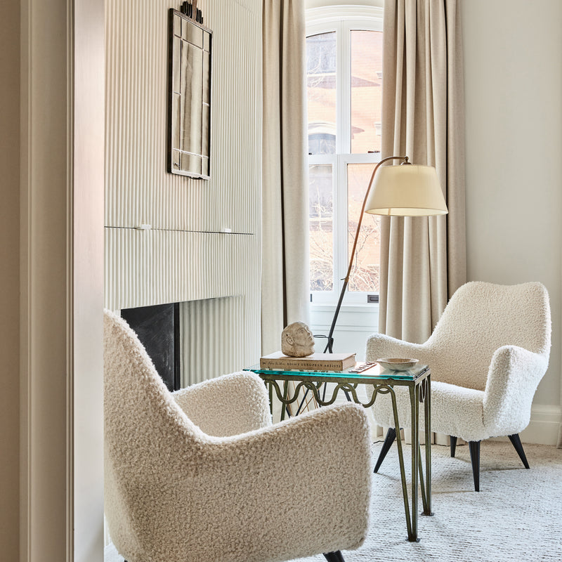 An inviting seating area in a primary bedroom consisting of two boucle reading chairs and a small table surrounding a fireplace. Designed by Nate Berkus and Jeremiah Brent. 