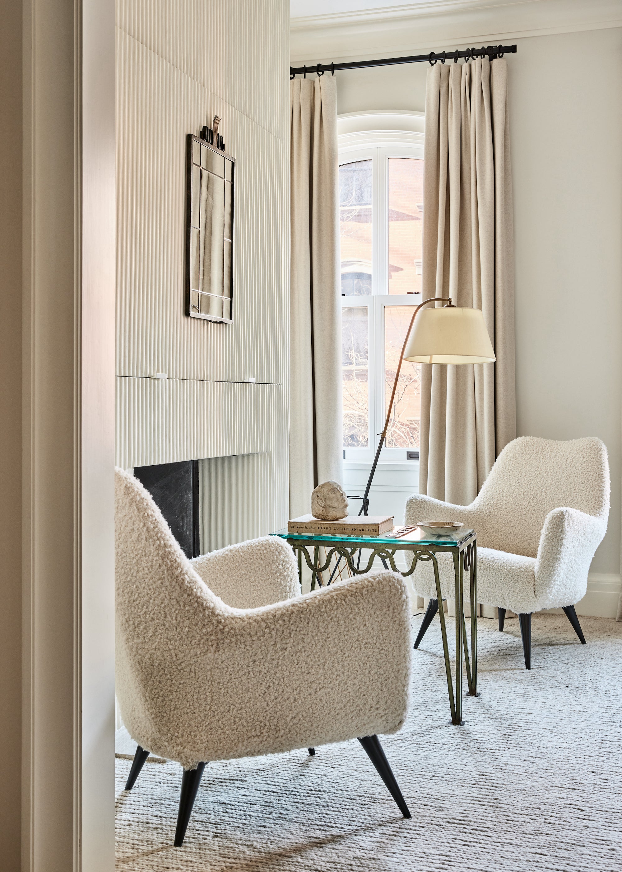 An inviting seating area in a primary bedroom consisting of two boucle reading chairs and a small table surrounding a fireplace. Designed by Nate Berkus and Jeremiah Brent. 