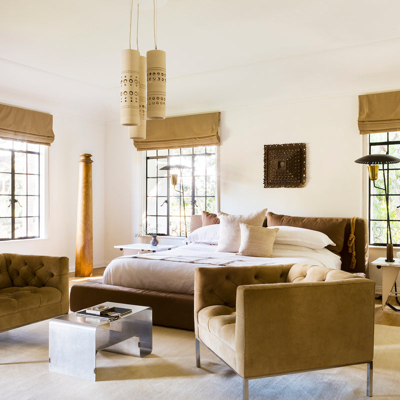 An open and airy primary bedroom, designed by Nate Berkus and Jeremiah Brent, with artful lighting, an upholstered platform bed, and a sitting area of two reading chairs and a sculptural table.