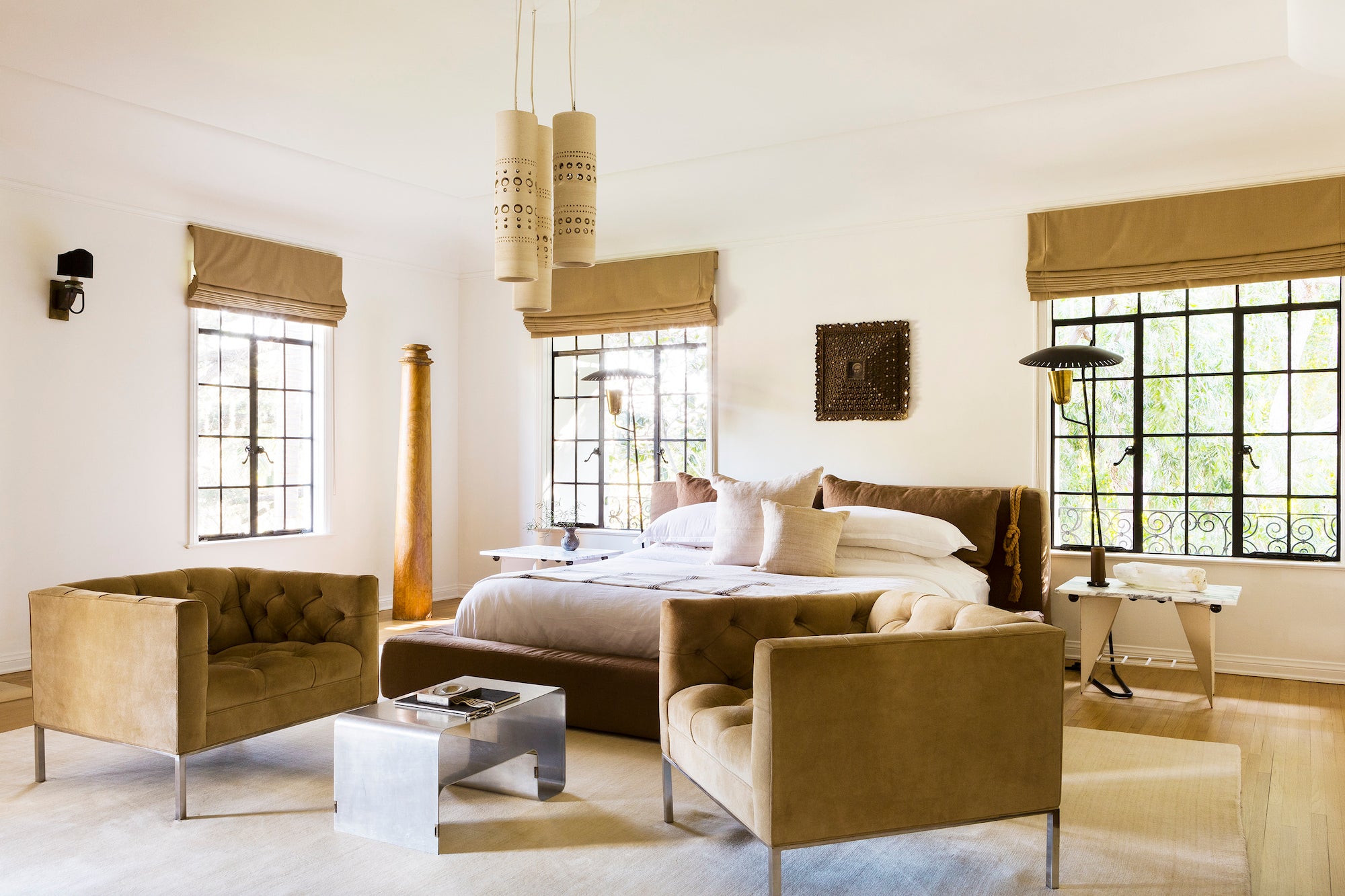 An open and airy primary bedroom, designed by Nate Berkus and Jeremiah Brent, with artful lighting, an upholstered platform bed, and a sitting area of two reading chairs and a sculptural table.